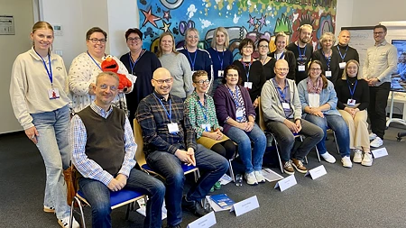 Gruppe von 20 Personen in einem Seminarraum vor einem bunten Banner, einige sitzend, andere stehend, mit Namensschildern